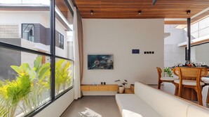 Ein modernes Wohnzimmer mit Sofa, Esstisch und Stühlen, einem großen Fenster mit Pflanzen und einem Gemälde an der Wand.