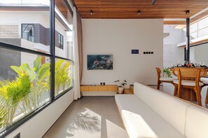 A modern living room with a sofa, dining table, and chairs, a large window with plants, and a painting on the wall. - Villa Kartal Altin by Pertama Stay (Badung Regency)