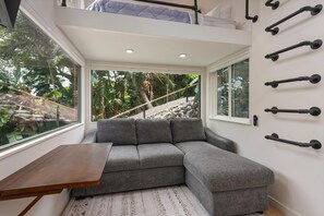 A compact living space with a grey sectional sofa, a small wooden table, and a view of greenery through large windows.