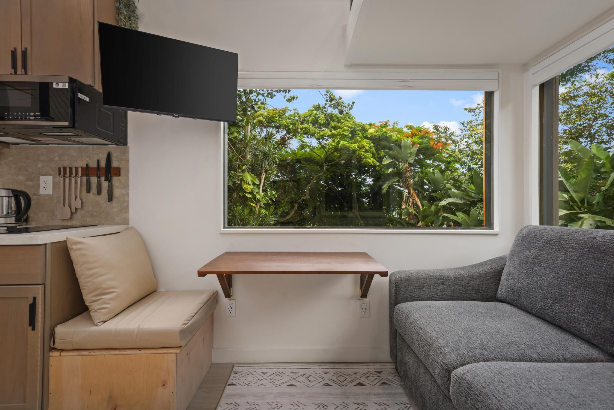A compact living space with a kitchenette, a sofa, a small table, and a window with a view of greenery.