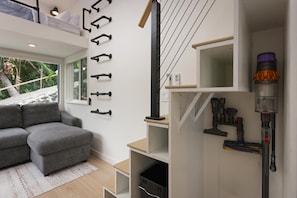 A modern living room with a grey sectional sofa, a white wall with black hooks, a window with a view of greenery, and a built-in shelf with cleaning tools.