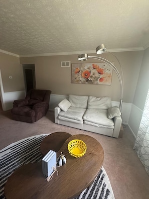 Ein Wohnzimmer mit einem braunen Sessel, einem weißen Sofa, einem runden Couchtisch mit einem gelben Korb und einem Bild an der Wand.