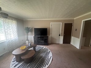 Ein Wohnzimmer mit Flachbildfernseher, rundem Couchtisch, Blumentopf und gemustertem Teppich.