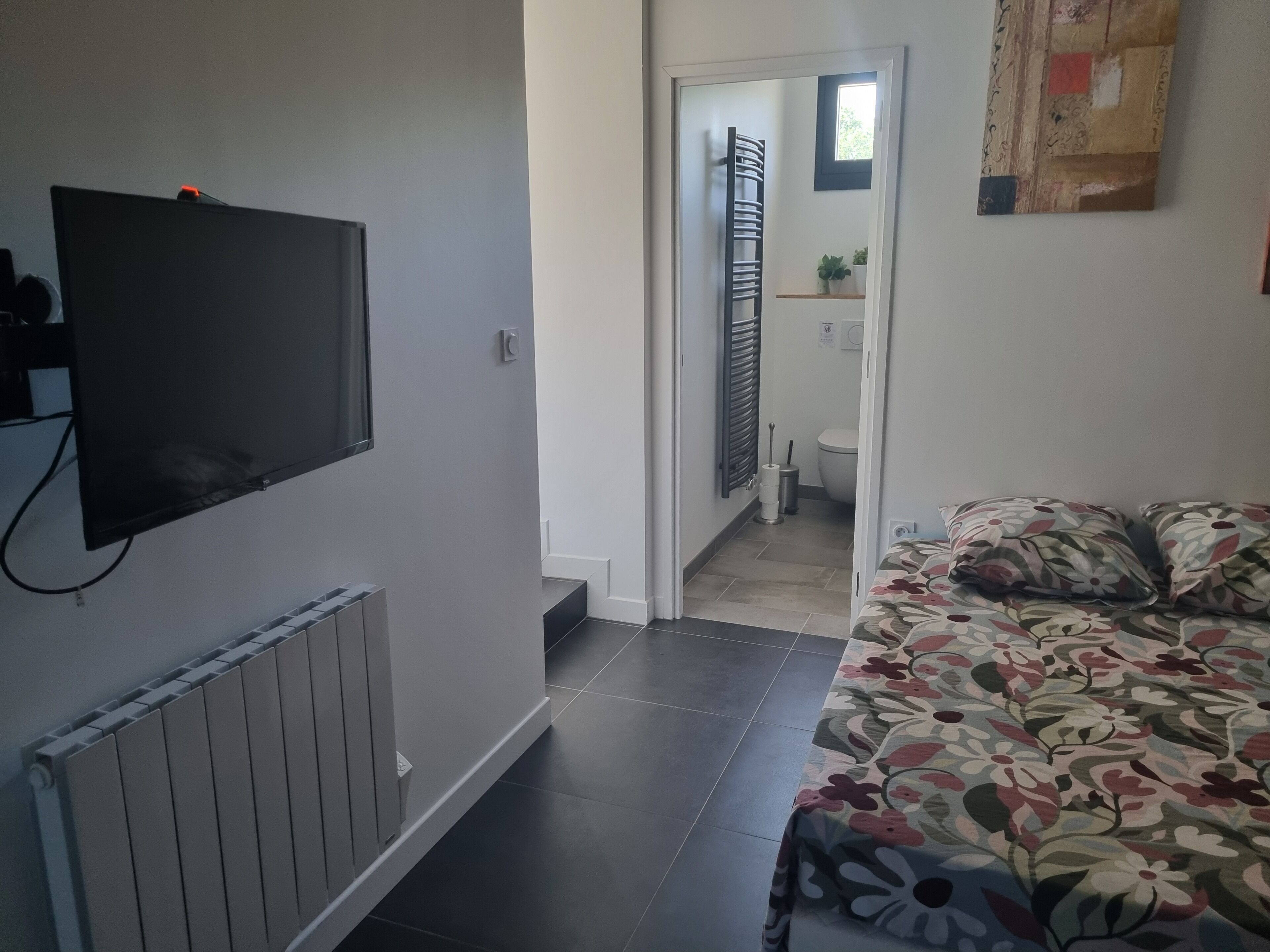 Een slaapkamer met een bed, een radiator, een televisie en een zichtbare badkamer door een open deur.