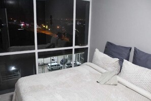 A hotel room with a large window, a bed with white bedding, and a view of a parking lot and city lights.