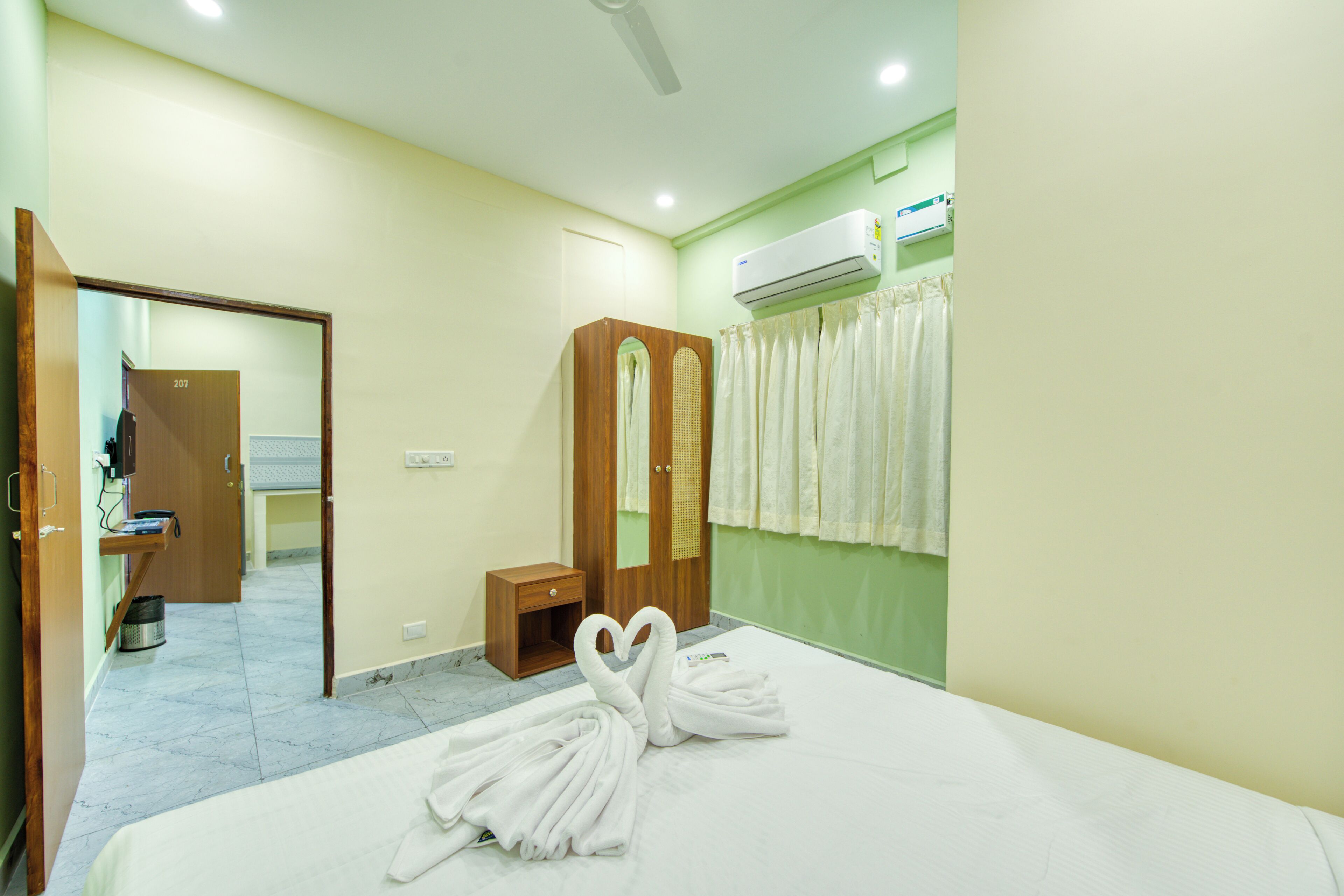 A hotel room with a bed, a wooden wardrobe, a small bedside table, a mirror, an air conditioning unit, and a bathroom with a shower curtain.