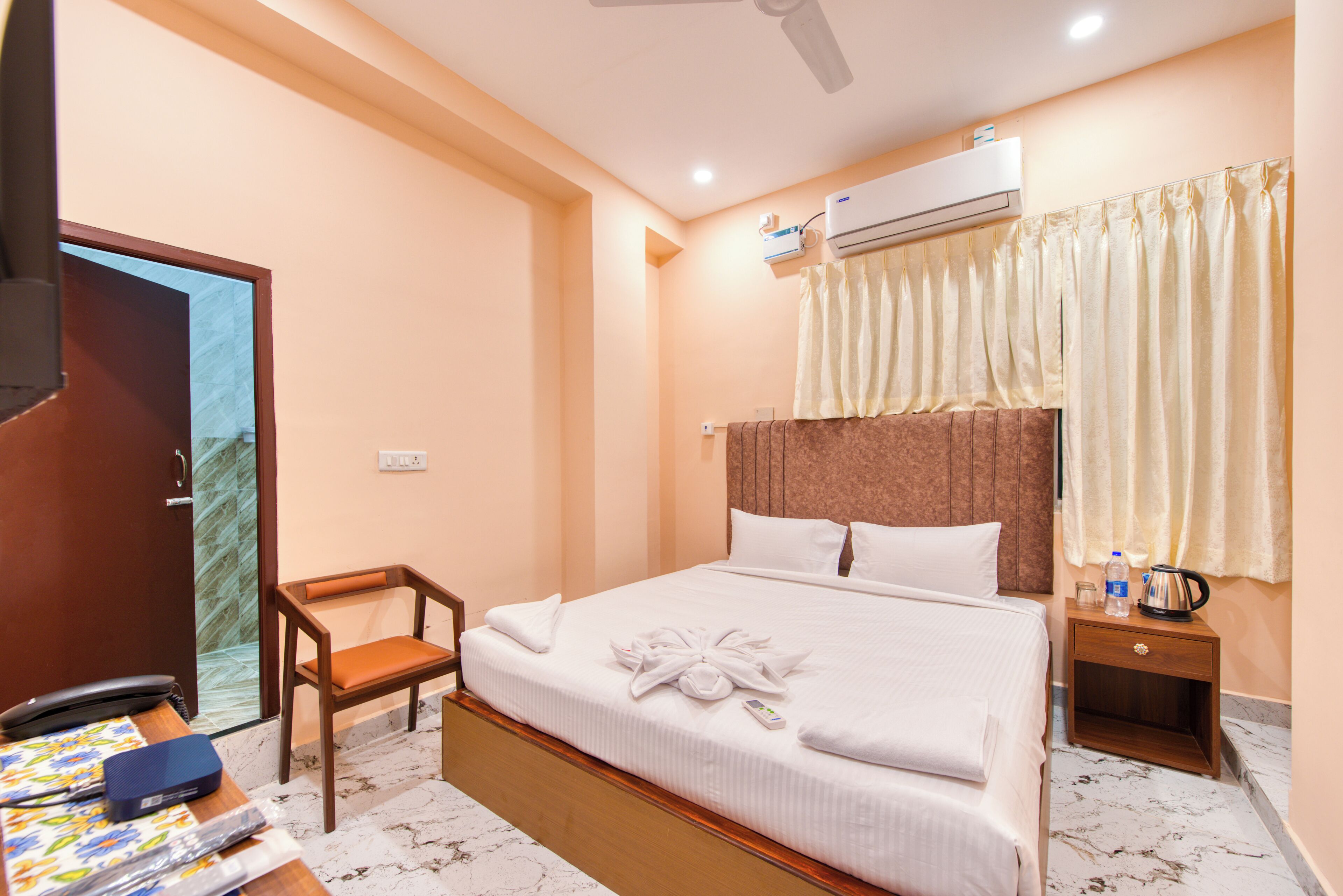A hotel room with a bed, a chair, a bedside table with a kettle, and an air conditioning unit.