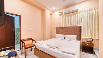 A hotel room with a bed, a chair, a bedside table with a kettle, and an air conditioning unit.