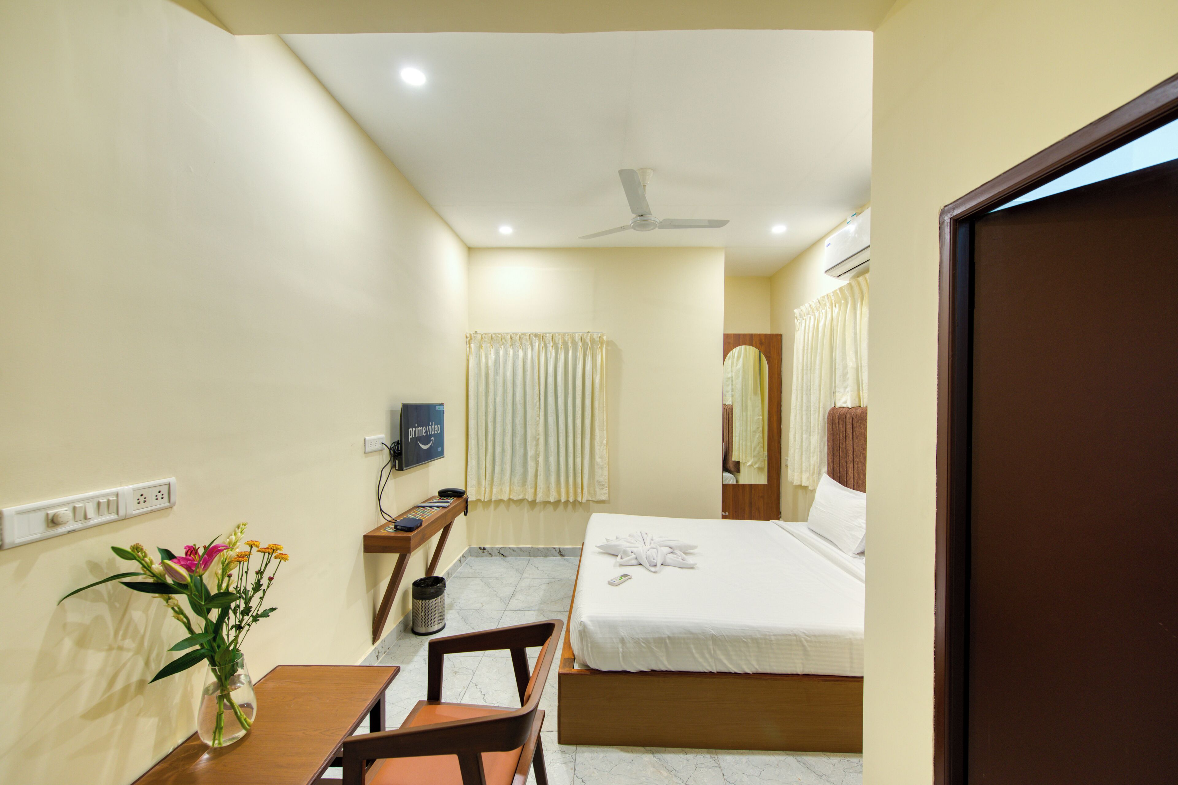 Quarto de hotel com cama, escrivaninha com cadeira, vaso com flores, televisão na parede e ventoinha no teto.