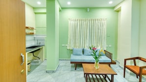 Una habitación con sofá, una mesita con un florero, una silla de madera, una zona de cocina con fregadero y una ventana con cortinas.