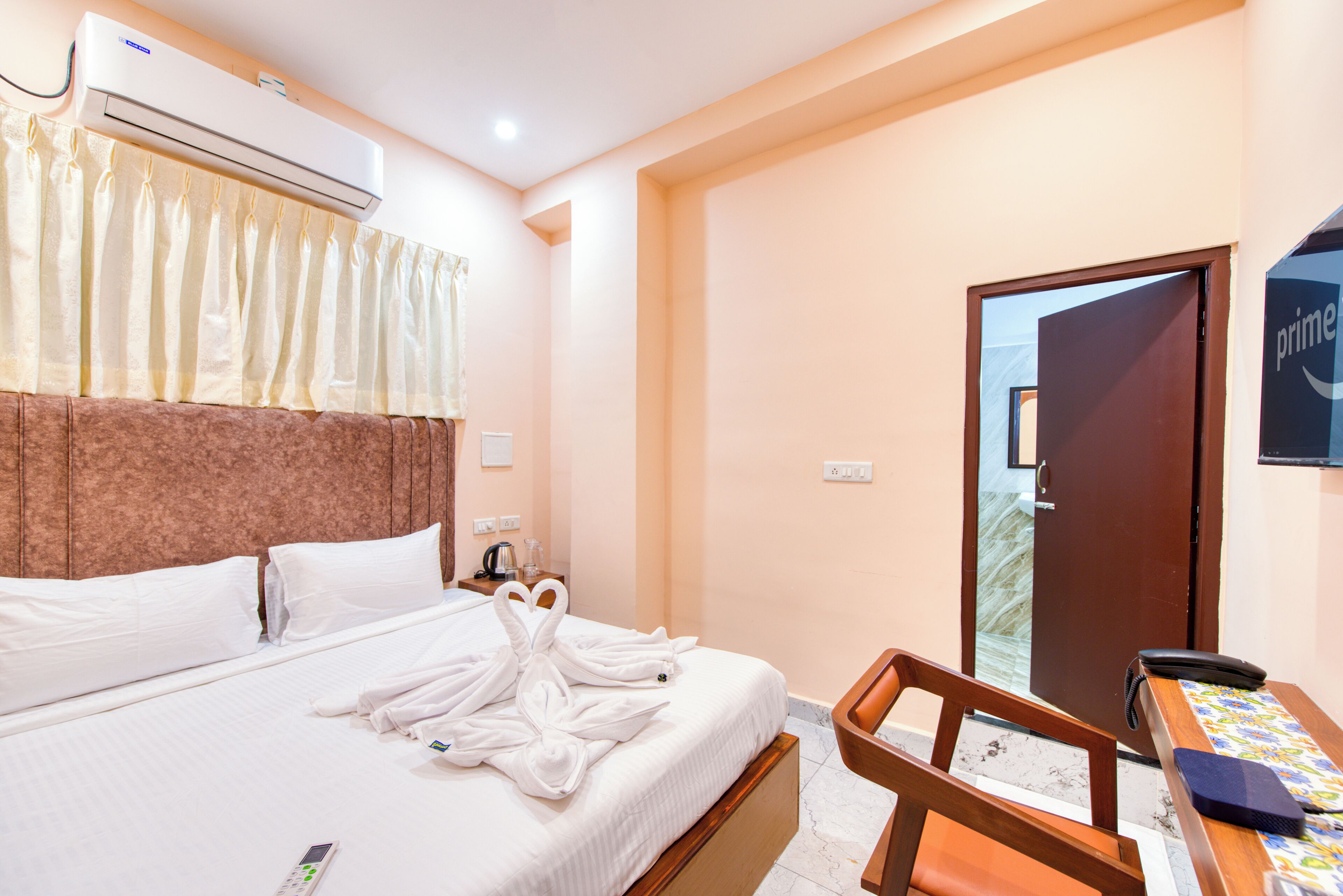 A hotel room with a bed, a desk with a telephone, a window with curtains, and an air conditioning unit.