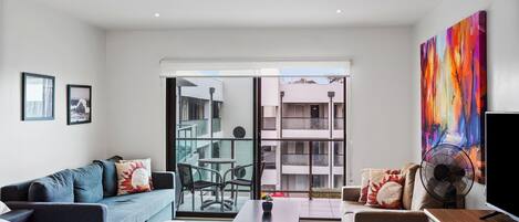 Una sala de estar moderna con un sofá, una mesa de centro y un televisor. Hay un balcón con puerta de vidrio que da a un espacio exterior.