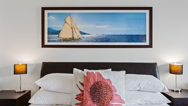 Une chambre d’hôtel avec un lit, deux lampes de chevet, un tableau encadré représentant un voilier et un coussin décoratif.