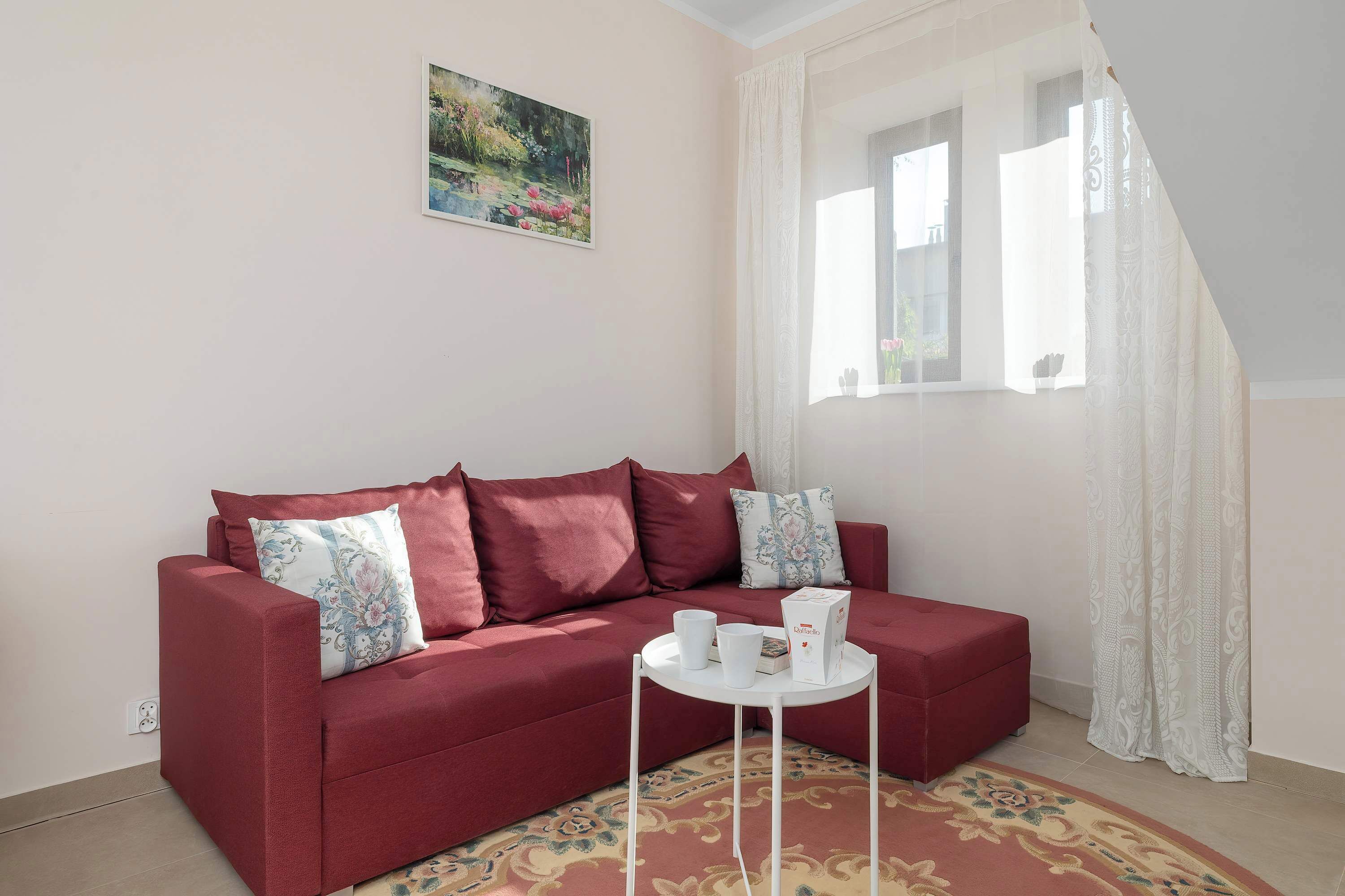 Un divano rosso con cuscini floreali, un tavolino bianco con una scatola e due tazze, un tappeto decorato e un quadro appeso al muro.
