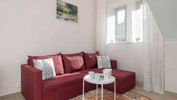 Un divano rosso con cuscini floreali, un tavolino bianco con una scatola e due tazze, un tappeto decorato e un quadro appeso al muro.