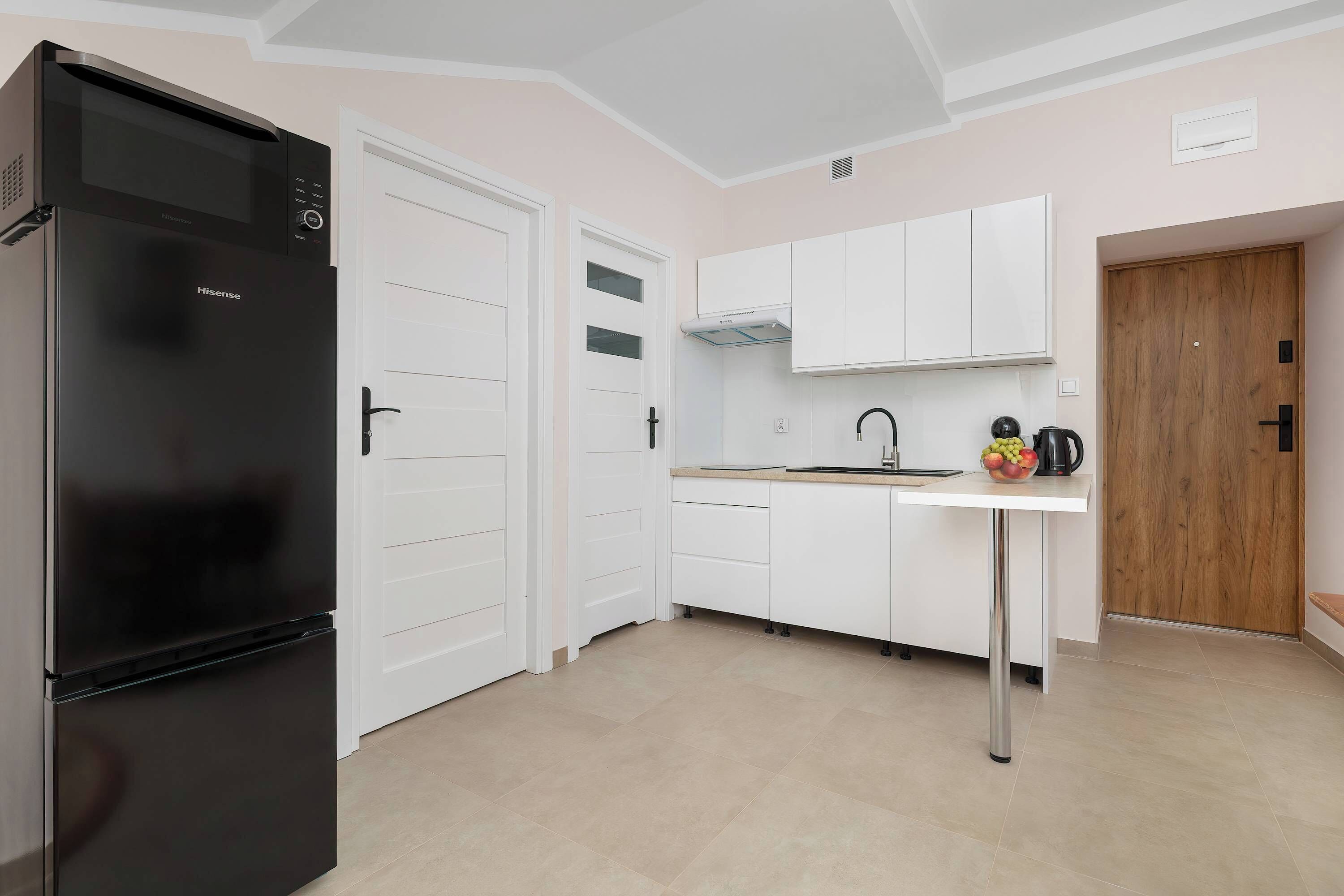 Une cuisine avec un réfrigérateur noir, des placards blancs, une porte en bois et un évier avec une bouilloire et un panier de fruits.