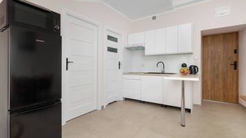 Une cuisine avec un réfrigérateur noir, des placards blancs, une porte en bois et un évier avec une bouilloire et un panier de fruits.