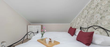 Un dormitorio con tragaluz, una cama con ropa blanca, almohadas rojas, una mesita de noche con un jarrón de flores y una bandeja con tazas y un plato.
