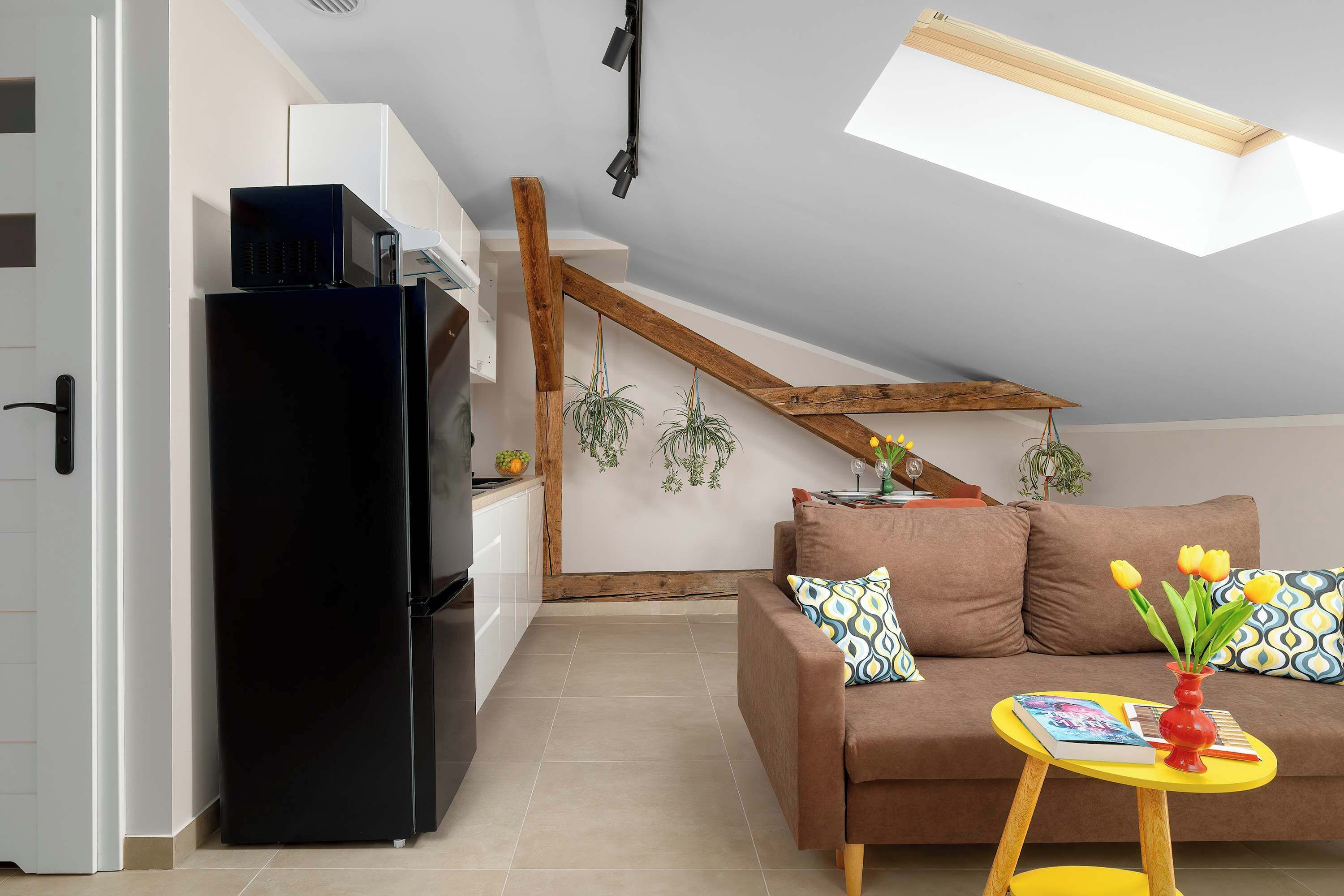 A modern kitchen with a black refrigerator, a wooden ceiling beam, and a dining area with a brown sofa and a yellow table.