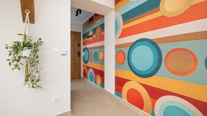 Um corredor com um mural colorido e abstrato na parede, uma planta pendente e um vigamento de madeira no teto.