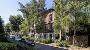 Uma rua residencial com carros estacionados, árvores e um edifício de dois andares com fachada de tijolos.