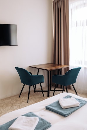 Una habitación de hotel con una cama, dos sillas color turquesa, una mesa de madera, un televisor montado en la pared y un ventanal con cortinas.