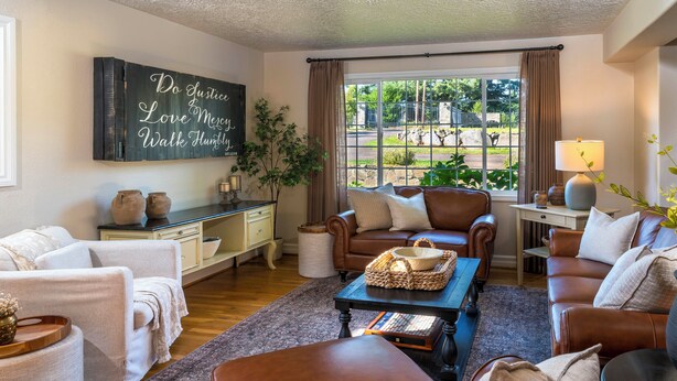 A cozy living room with a brown leather sofa, a coffee table with a woven basket, a large window with a view of greenery, and a wall-mounted sign with a motivational quote. - Luxe Home & Glamping w/ Pond in Oregon City! (Oregon City)