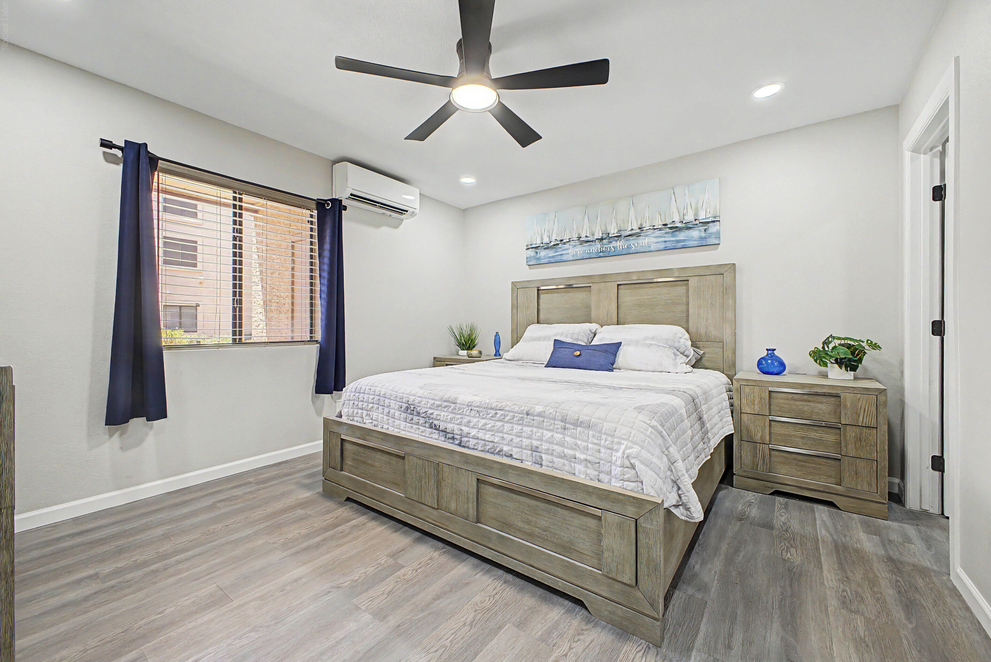 A bedroom with a bed, nightstands, a ceiling fan, an air conditioning unit, a window with curtains, and a painting on the wall.