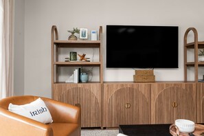 A living room with a leather armchair, a television on the wall, wooden shelves with decorative items, and a cabinet with a flat-screen TV. - Walk to ND Luxury 2BR/2BA Condo Balcony + Parking (South Bend)