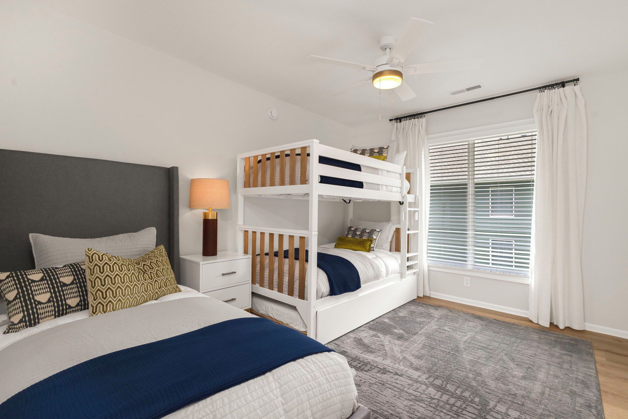 Une chambre avec un lit superposé, un lit simple, une table de chevet avec une lampe, une fenêtre avec des stores et un ventilateur de plafond.