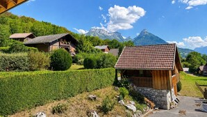 A wooden cabin with a stone foundation, surrounded by a well-maintained garden and a mountain backdrop.