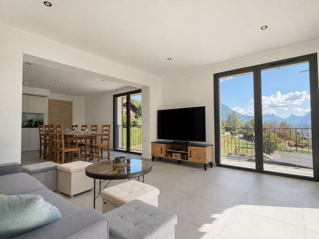 A modern living room with a flat-screen TV, a dining area with wooden furniture, and large windows offering a mountain view.