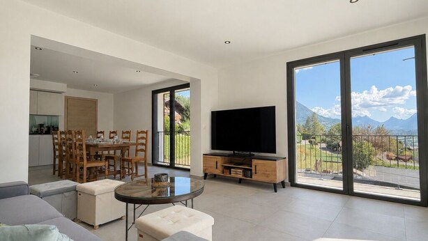 A modern living room with a flat-screen TV, a dining area with wooden furniture, and large windows offering a mountain view.
