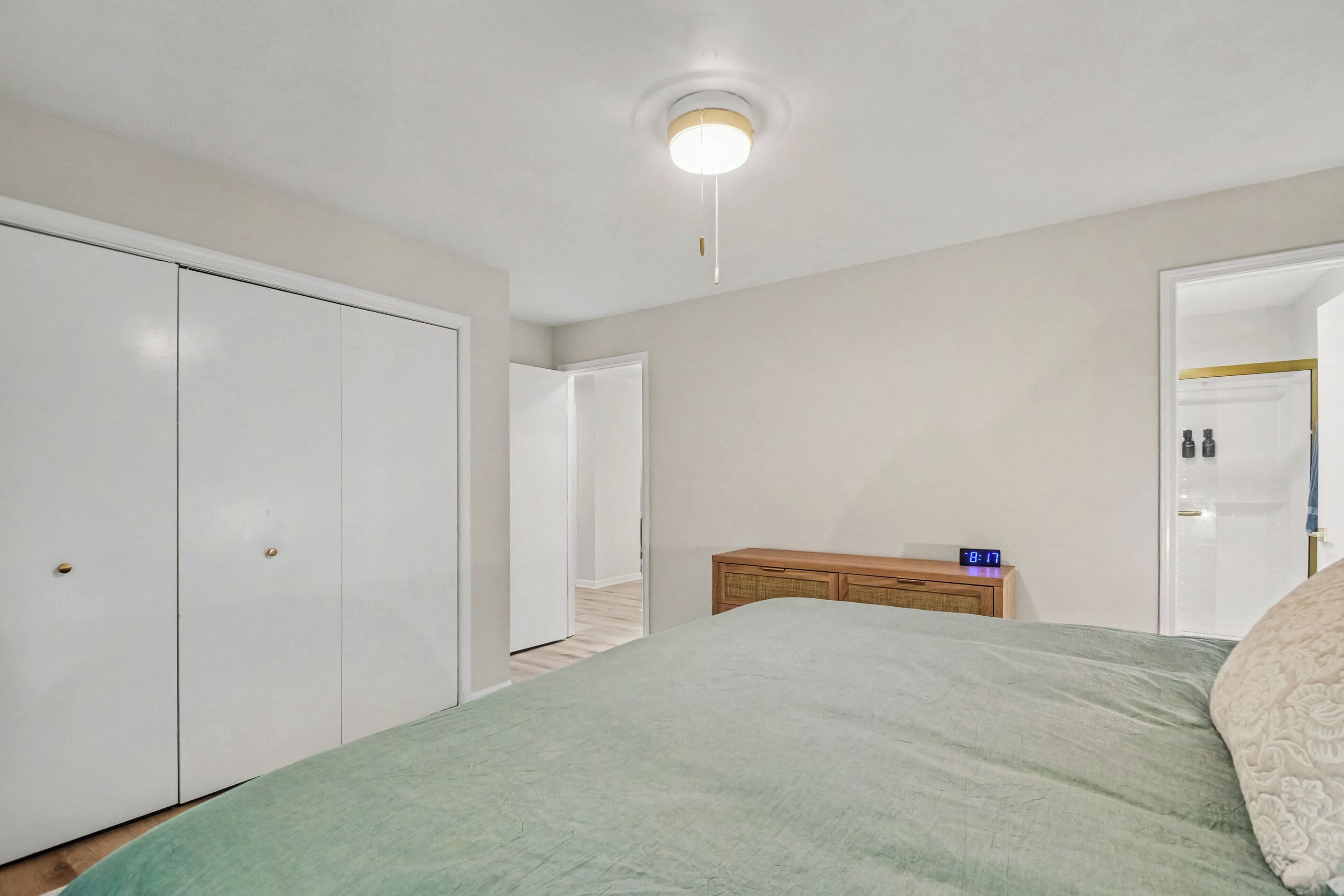 A bedroom with a bed, a bedside table with a clock, a wardrobe, and a door leading to another room.
