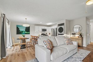 Ein modernes Wohnzimmer mit einem grauen Sofa, Holzstühlen, einem Esstisch und einer Küche im Hintergrund.