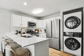 Cocina moderna con armarios blancos, electrodomésticos de acero inoxidable, una isla central con taburetes de bar y lavadora y secadora integradas.