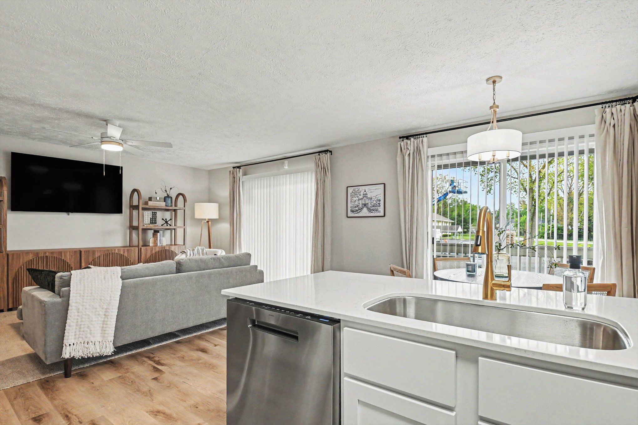 A modern kitchen with a dishwasher, a dining area with a table and chairs, a gray sofa, and a TV mounted on the wall.