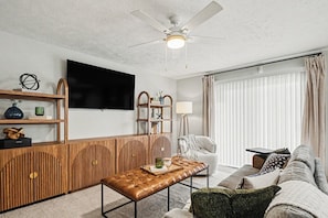 A living room with a flat-screen TV, wooden furniture, a ceiling fan, and a window with blinds.