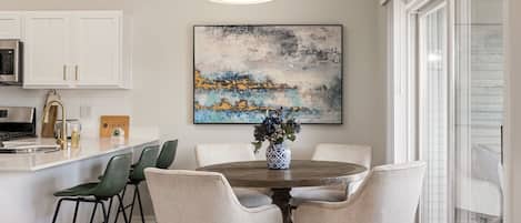 A dining area with a round table, six chairs, a vase with flowers, a painting on the wall, and a kitchen in the background.