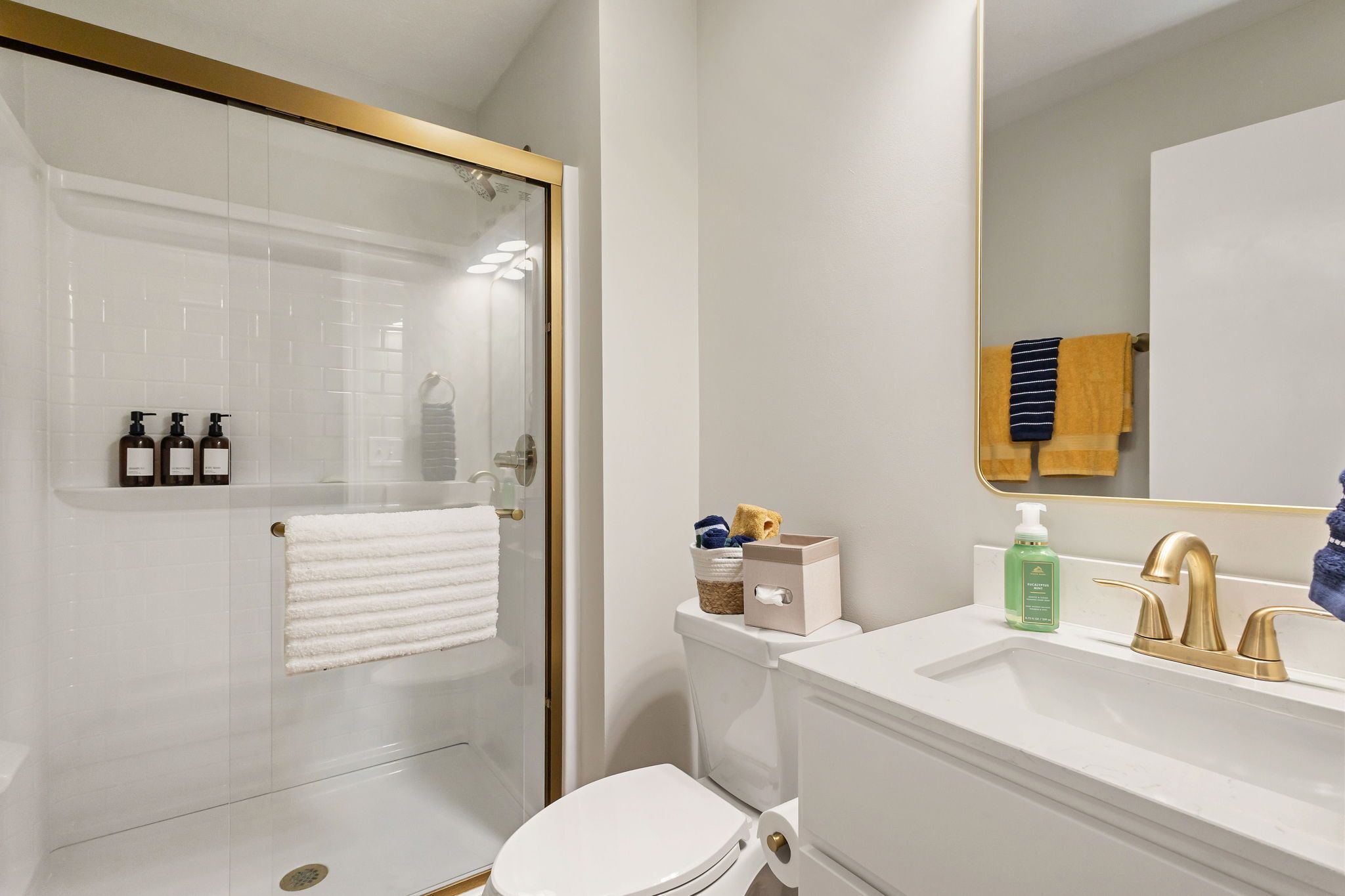 Ein modernes Badezimmer mit einer gläsernen Duschkabine, einer weißen Toilette, einem Waschbecken mit goldenen Armaturen und einem Spiegel mit einem daran hängenden Handtuch.