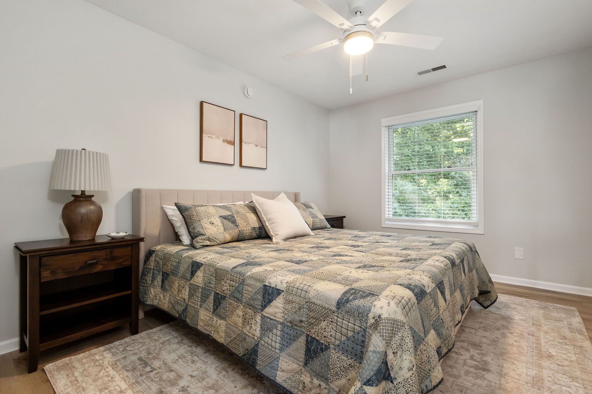 A bedroom with a bed, a nightstand, a lamp, a ceiling fan, and a window with blinds.