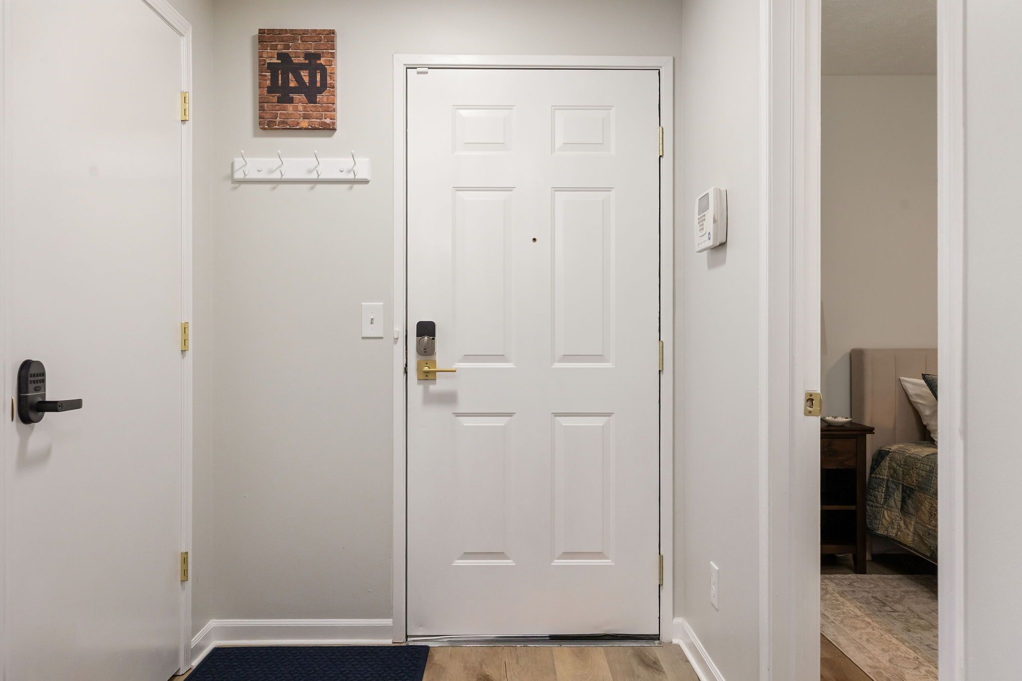 Un couloir avec une porte blanche, un porte-manteau, un thermostat et un lit à l’arrière-plan.