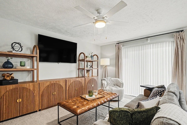 A living room with a flat-screen TV, wooden furniture, a ceiling fan, and a window with blinds.