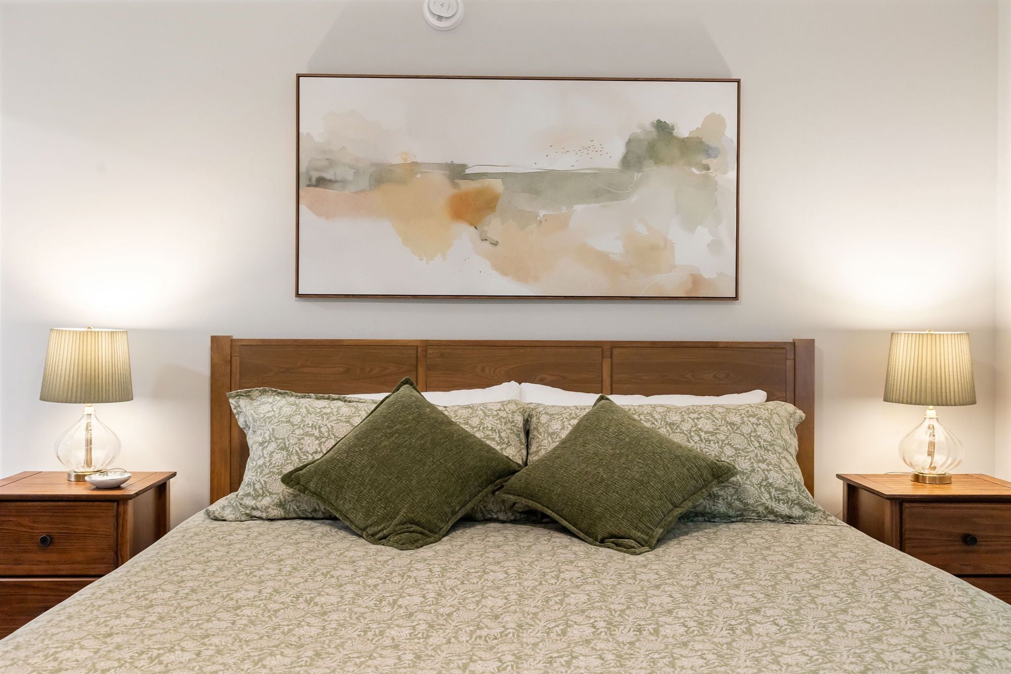 A bed with a patterned bedspread, two green pillows, and two bedside lamps on wooden nightstands.