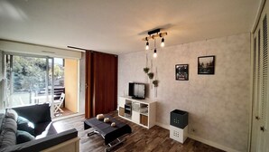 A modern living room with a sofa, a coffee table, a TV on a white cabinet, and a sliding glass door leading to an outdoor area. - Studio le banga (Ax-les-Thermes)