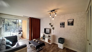Een moderne woonkamer met een bank, een salontafel, een televisie op een witte kast, en een schuifdeur die leidt naar een buitenruimte.