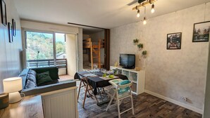 A cozy living area with a sofa, dining table, and TV. There is a balcony with a view of trees. - Studio le banga (Ax-les-Thermes)