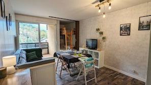 Een gezellige woonkamer met een bank, eettafel en televisie. Er is een balkon met uitzicht op bomen.