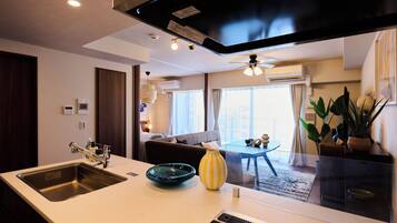 Quarto de hotel moderno com cozinha, área de estar com sofá e mesa de jantar, e vista para o horizonte urbano por grandes janelas.