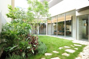 Un espacio exterior moderno con puerta de cristal que da a un área de descanso, rodeado de abundante vegetación y un camino de piedra con diseño.